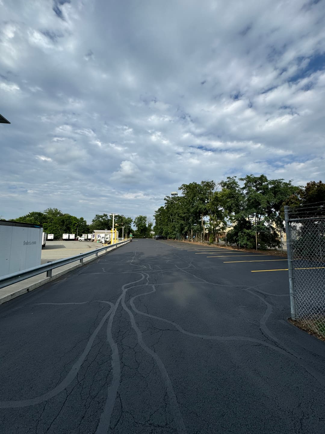 Empty asphalt road with scattered trees under a cloudy sky. Industrial area in the background.