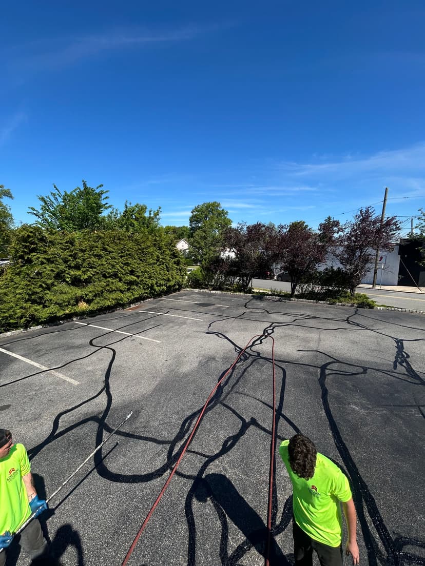 Workers applying sealant on a parking lot with vibrant green trees under a clear blue sky.