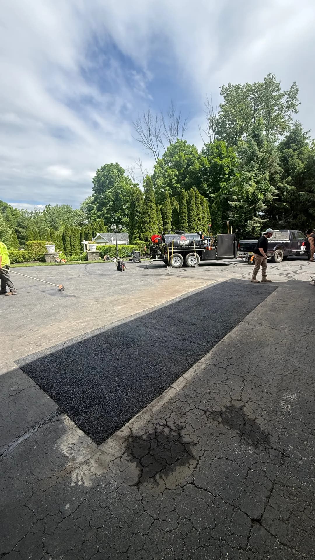 Asphalt paving crew working on a driveway with machinery and trees in the background.