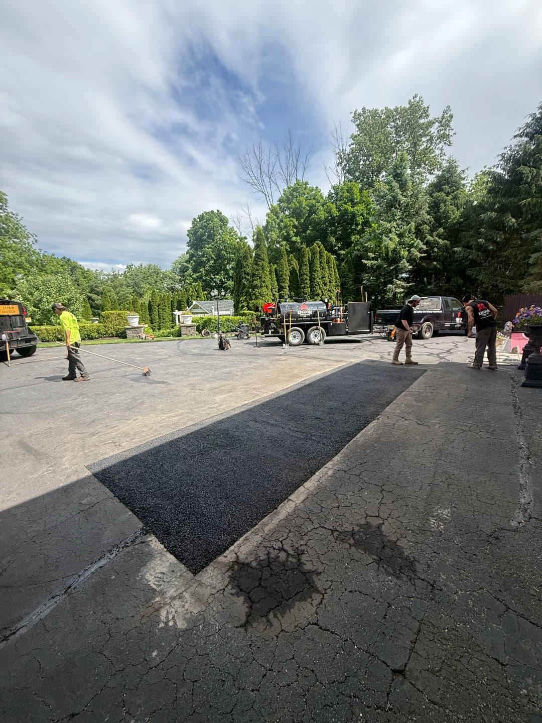 Workers laying asphalt on a driveway with trees and vehicles in the background.