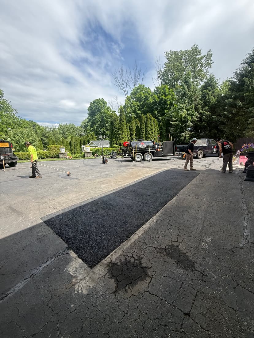 Workers laying asphalt on a driveway with trees and vehicles in the background.