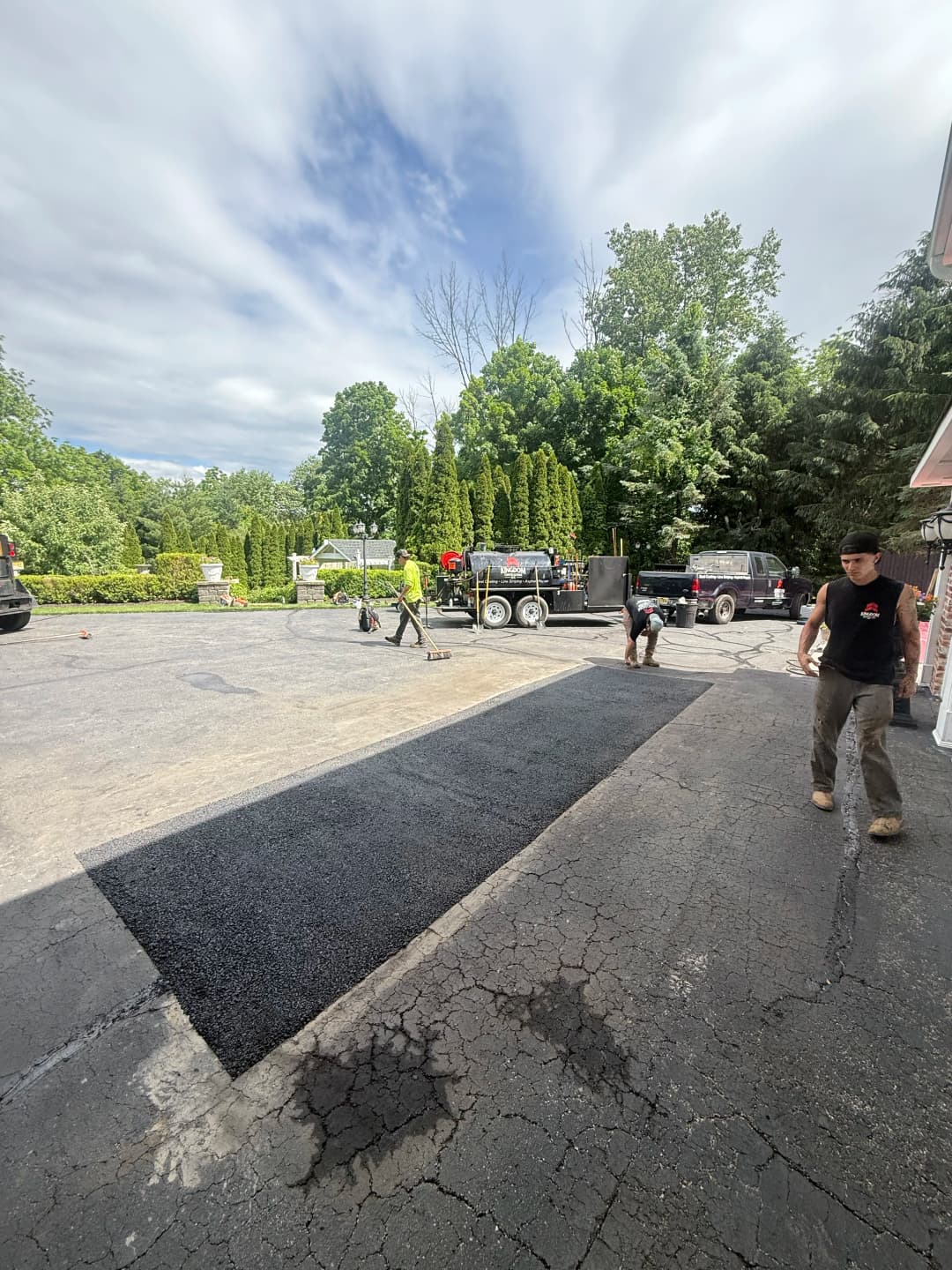 Workers laying asphalt on a driveway with construction vehicles and greenery in the background.