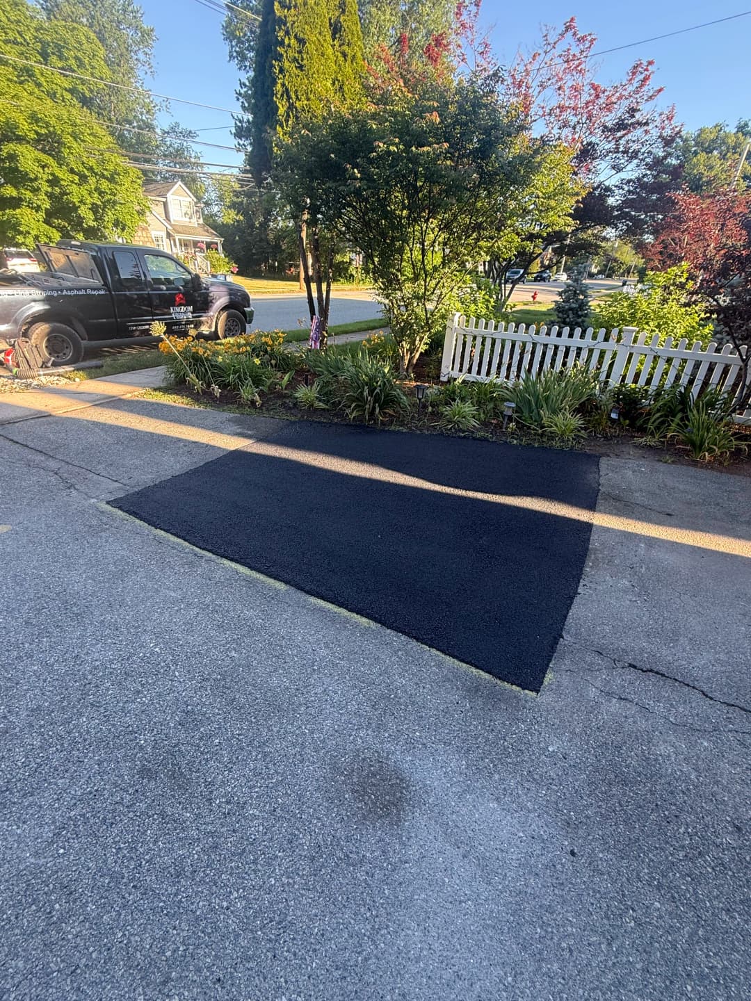Freshly paved blacktop driveway next to a garden and white picket fence on a sunny day.