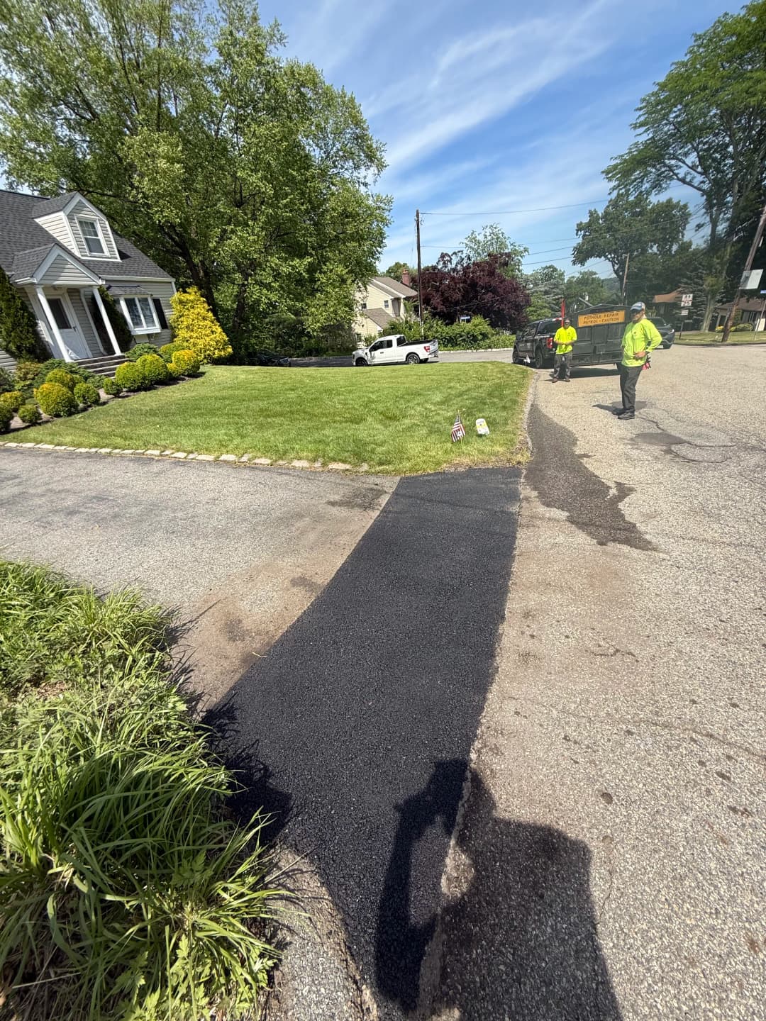 Workers applying asphalt to a roadway near a residential home on a sunny day.