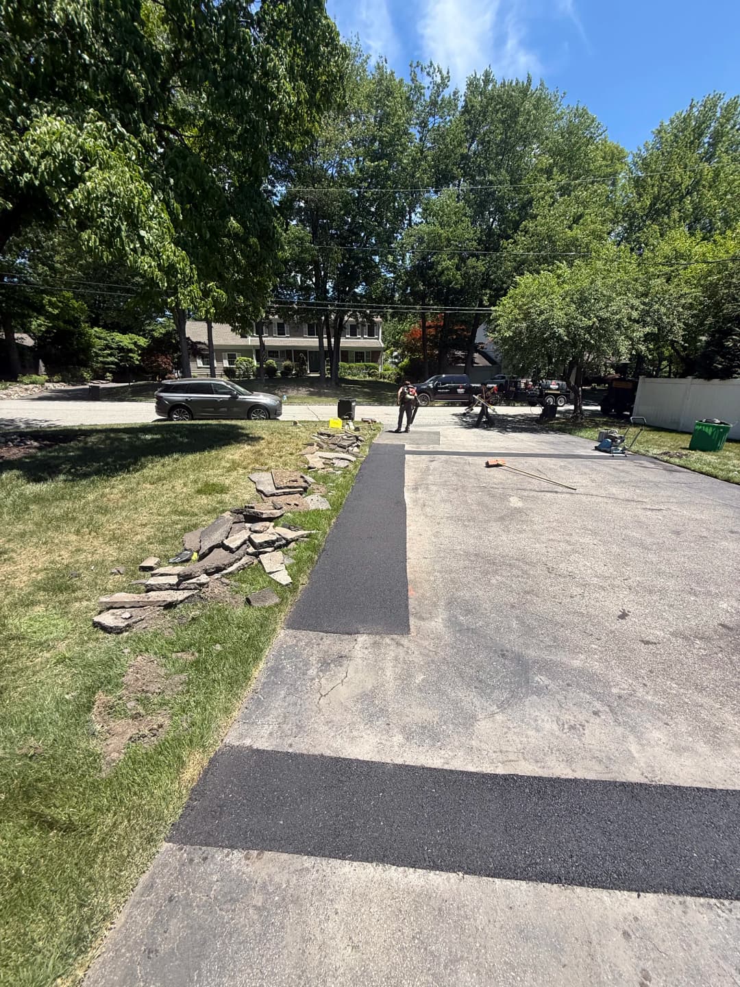 Person applying asphalt on a driveway with trees and a house in the background.
