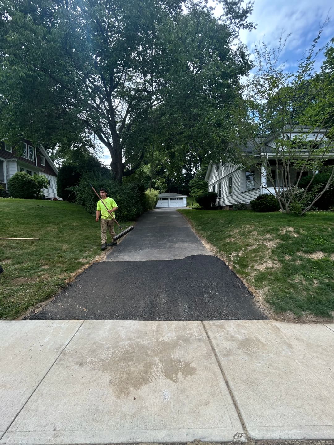 Worker smoothing freshly paved driveway beside residential homes and greenery.