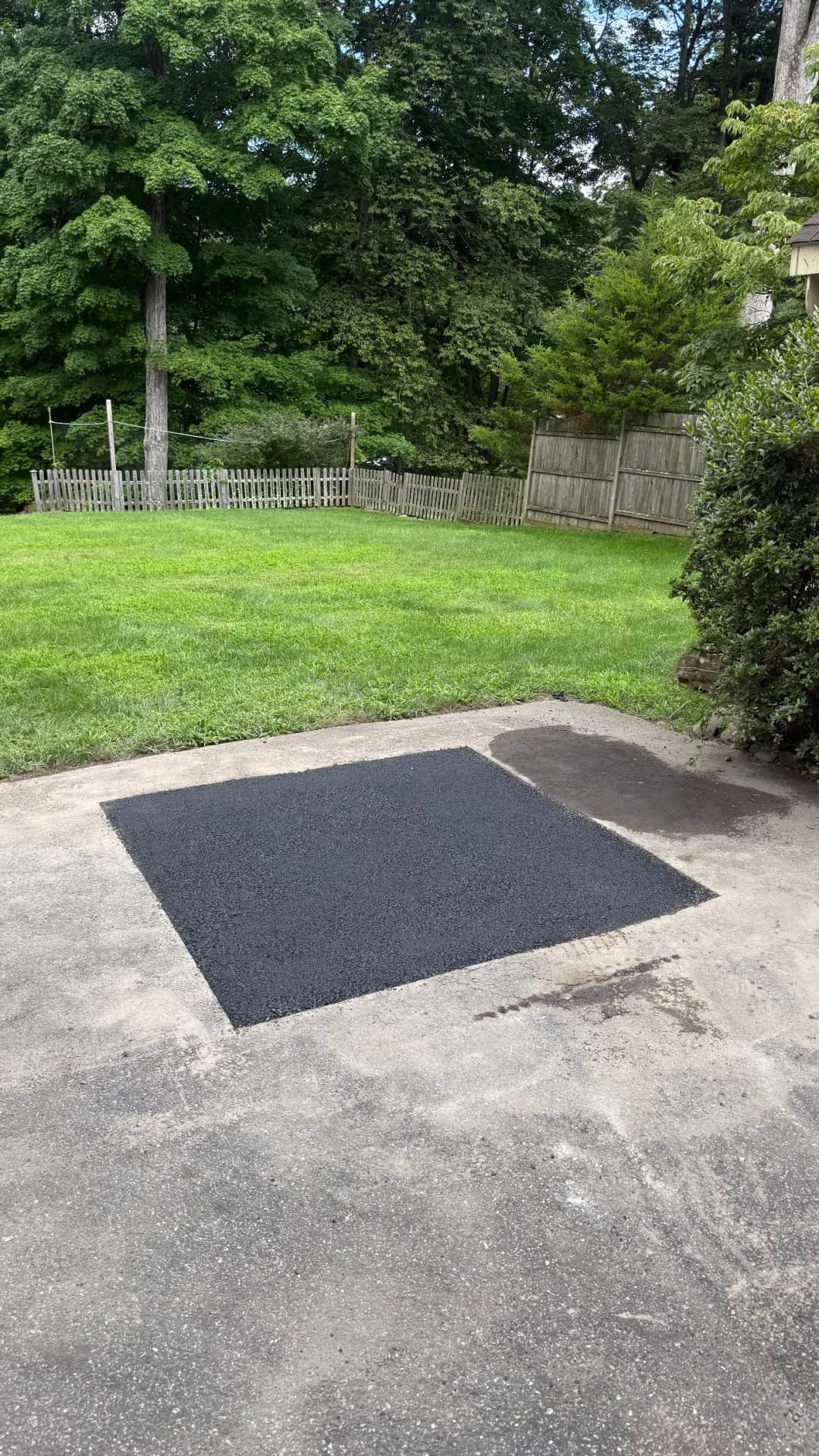 Freshly paved black asphalt patch on a driveway in a grassy yard with wooden fence.