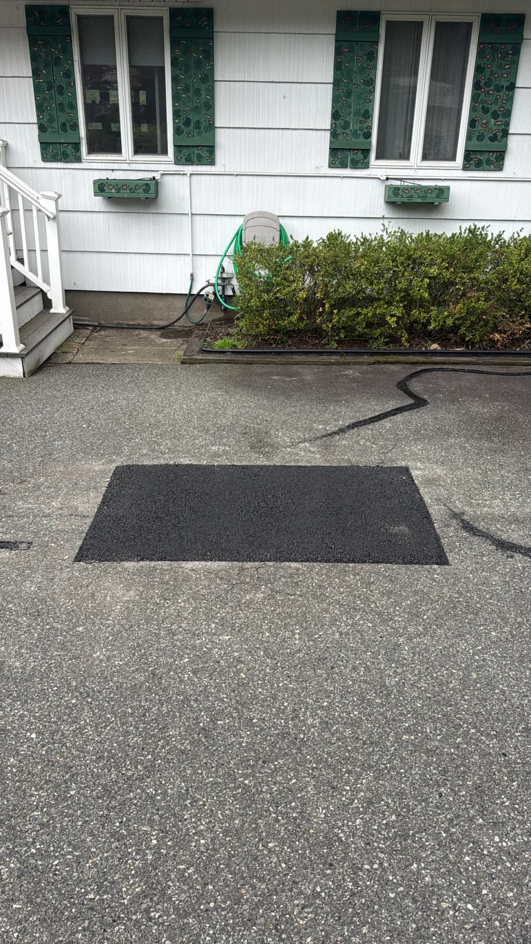 Black asphalt patch on a driveway with white house and greenery in the background.