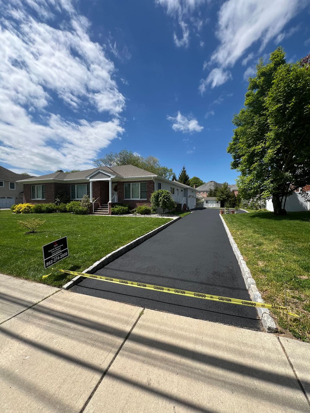 Newly paved driveway with caution tape in front of a brick house under a blue sky.
