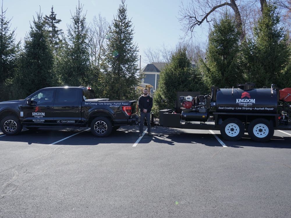 Kingdom seal coating truck and trailer parked, worker standing in front with trees in background.