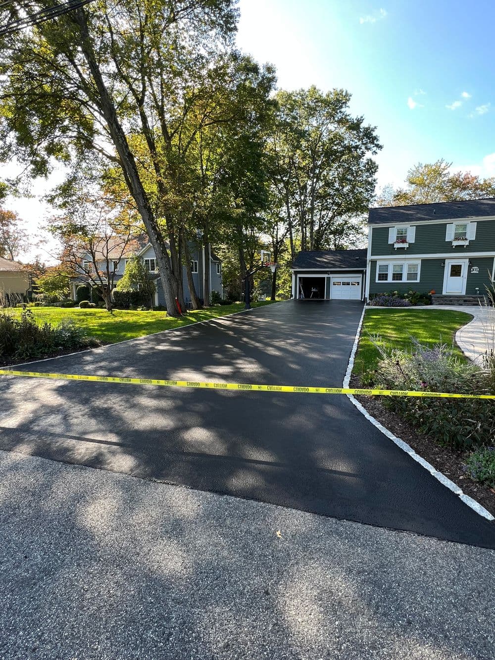 Freshly paved driveway with caution tape, surrounded by trees and houses under clear blue sky.