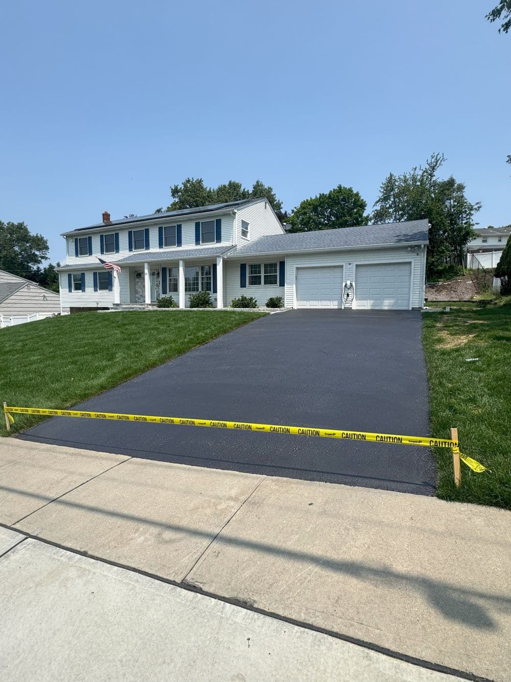 Freshly sealed asphalt driveway with caution tape near a white two-story house.