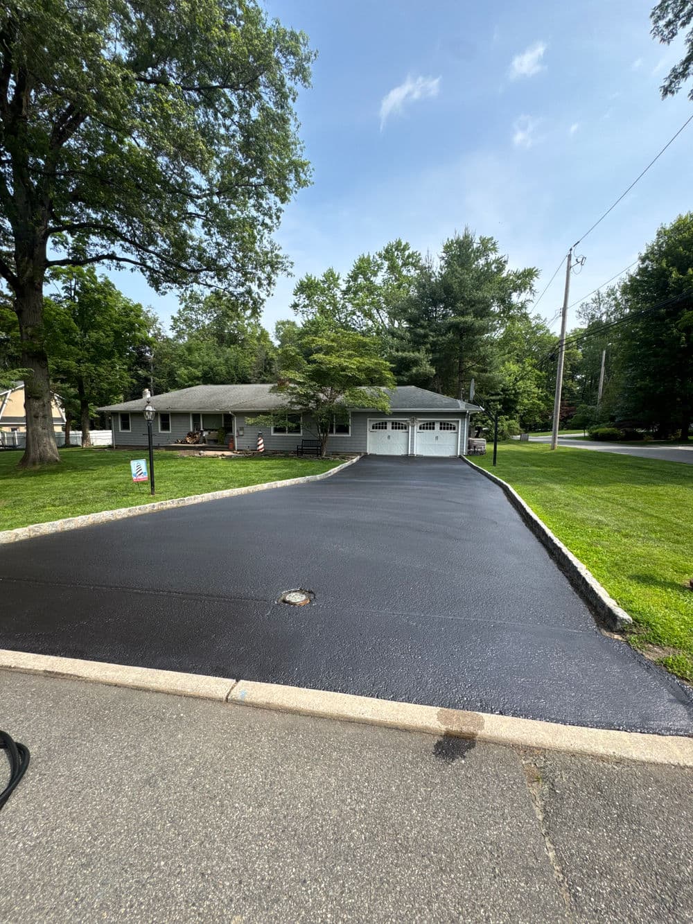 Freshly paved driveway leading to a gray house surrounded by greenery on a sunny day.