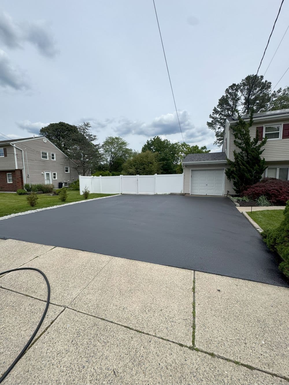 Freshly sealed blacktop driveway with white vinyl fence and suburban homes in the background.
