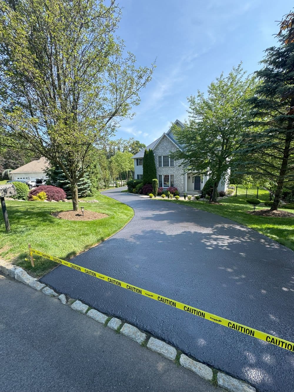 Home with freshly paved driveway and caution tape, surrounded by greenery and landscaped yard.