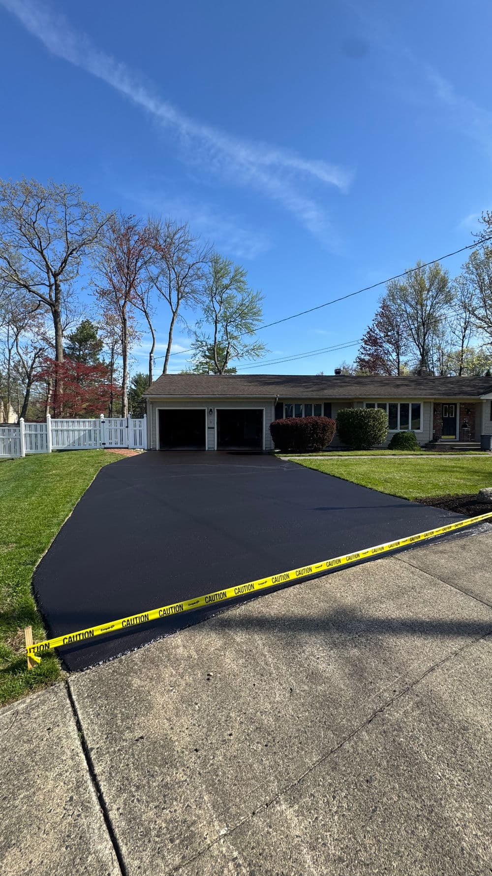 Freshly paved black asphalt driveway with caution tape and a suburban home in the background.