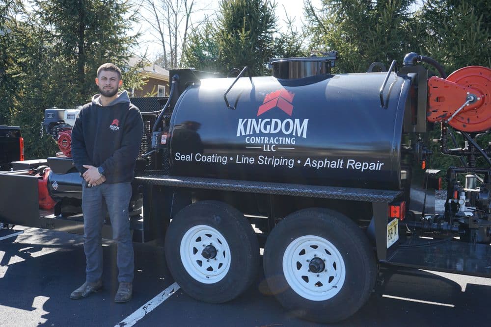 Man standing next to a Kingdom Contracting LLC seal coating truck in a wooded area.