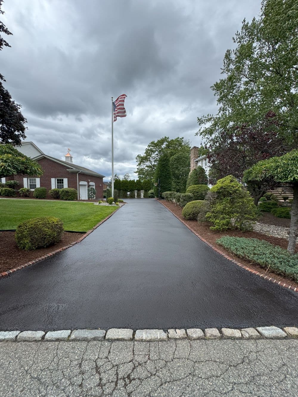 Paved driveway leads to a house with lush landscaping and an American flag under cloudy skies.