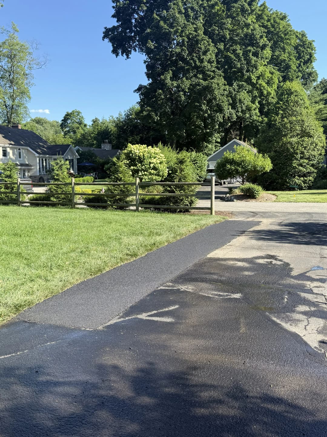 Freshly paved asphalt driveway leading to homes with lush greenery and a blue sky.