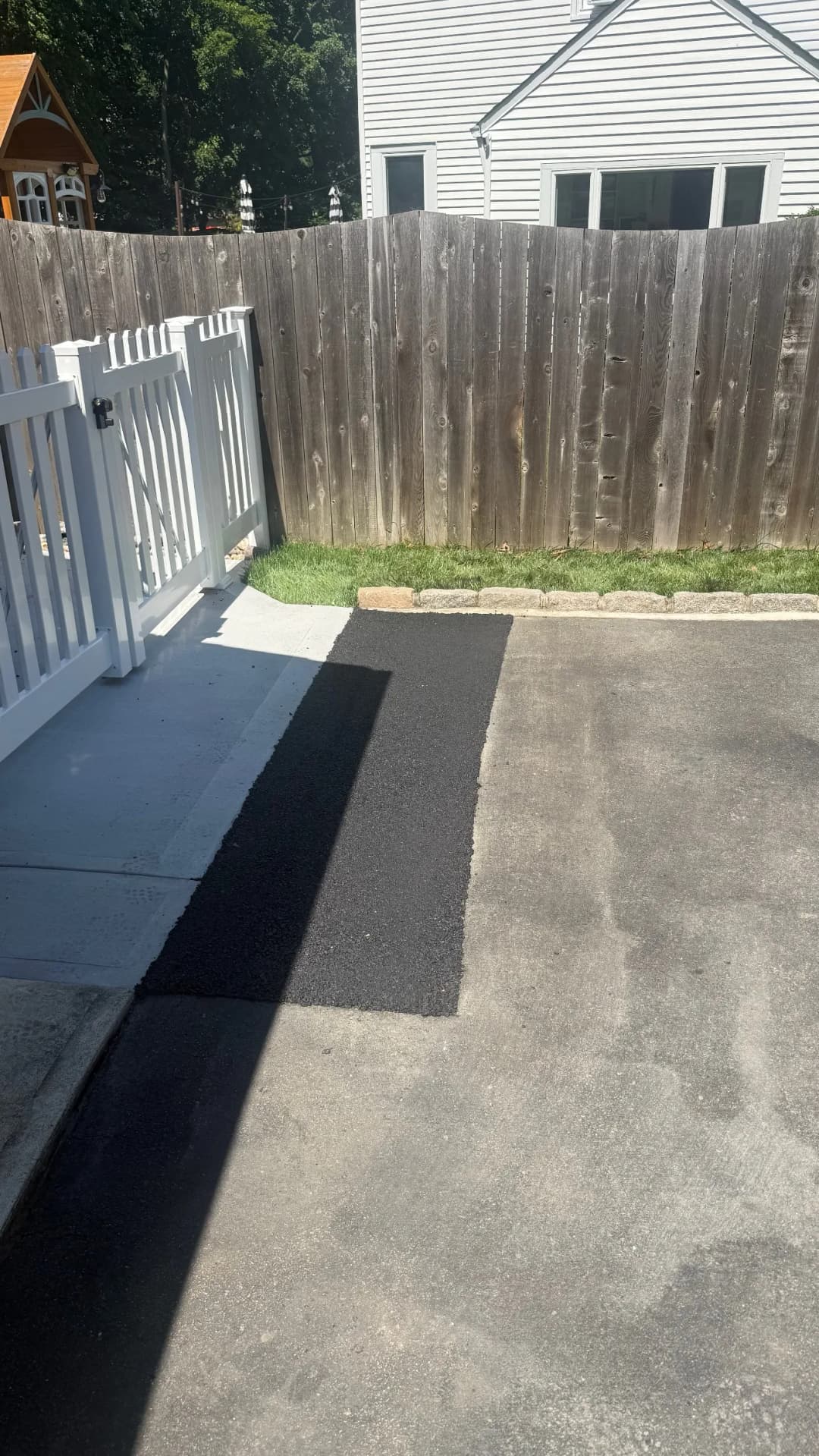 Freshly paved black asphalt driveway next to white picket fence and wooden fence.